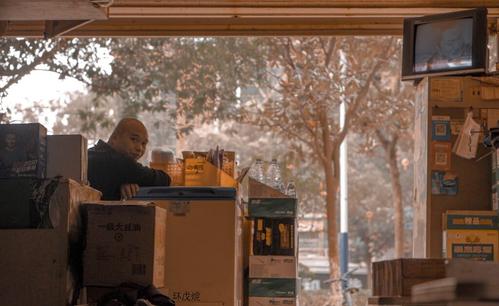 Person sits on the floor surrounded by cardboard moving boxes, looking thoughtfully overwhelmed in a bright, modern room, ...