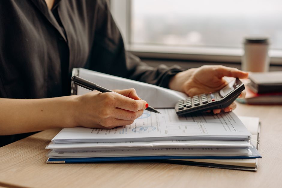 Professionally dressed person sits at a modern desk, looking concerned as they review a stack of official-looking document...